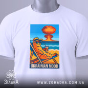 Футболка український настрій, біла, складена на столі, фон світло-сірий, принт з котом у сонцезахисних окулярах, що сидить на шезлонгу з напоєм, на фоні пляжу та вибуху, з етикеткою на комірці.