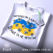 Футболка українці як бджоли, білого кольору, складена на поверхні, фон світло-сірий, принт з жовто-блакитними кольорами, бджолами та написом 'Краще їх не чіпати', з зеленим ярликом.