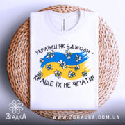 Футболка українці як бджоли, білого кольору, складена на плетеній кошику, фон світло-сірий, принт з жовто-блакитними кольорами, бджолами та написом 'Краще їх не чіпати'.