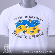 Футболка українці як бджоли, білого кольору, складена на поверхні, фон світло-сірий, принт з жовто-блакитними кольорами, бджолами та написом 'Краще їх не чіпати', з зеленим ярликом.