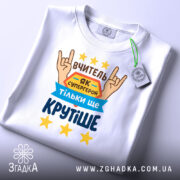 Футболка вчитель білого кольору, складена на столі, фон світло-сірий, принт з текстом "ВЧИТЕЛЬ ЯК СУПЕРГЕРОЙ ТІЛЬКИ ЩЕ КРУТІШЕ" та зображенням рук у жесті "рок", зірки жовтого кольору, на футболці висить етикетка.