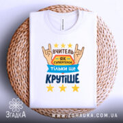 Футболка вчитель білого кольору, складена на плетеній основі, фон світло-сірий, принт містить текст "ВЧИТЕЛЬ ЯК СУПЕРГЕРОЙ ТІЛЬКИ ЩЕ КРУТІШЕ" з зображенням рук у жесті "рок" та жовтими зірками, видно етикетку на комірці.