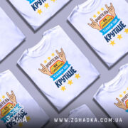 Футболка вчитель білого кольору, зображена з правого ракурсу на манекені, фон світло-сірий, принт містить текст "ВЧИТЕЛЬ ЯК СУПЕРГЕРОЙ ТІЛЬКИ ЩЕ КРУТІШЕ" з зображенням рук у жесті "рок", зірки жовтого кольору.