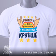 Футболка вчитель білого кольору, складена на столі, фон світло-сірий, принт з текстом "ВЧИТЕЛЬ ЯК СУПЕРГЕРОЙ ТІЛЬКИ ЩЕ КРУТІШЕ" та зображенням рук у жесті "рок", зірки жовтого кольору, на футболці висить етикетка.