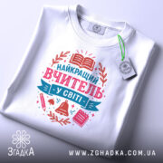 Футболка вчитель у світі білого кольору, складена на поверхні, фон світло-сірий, принт з текстом 'НАЙКРАЩИЙ ВЧИТЕЛЬ У СВІТІ' у рожевих та блакитних тонах, з елементами, такими як книга, дзвоник та ручка, з етикеткою на комірці.