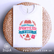 Футболка вчитель у світі білого кольору, складена на плетеній кошику, фон світлий, принт містить текст 'НАЙКРАЩИЙ ВЧИТЕЛЬ У СВІТІ' у рожевих та блакитних тонах, з елементами, такими як книга, дзвоник та ручка.