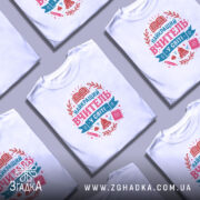 Футболка вчитель у світі білого кольору, зображена на манекені з правого боку, фон світло-сірий, принт містить текст 'НАЙКРАЩИЙ ВЧИТЕЛЬ У СВІТІ' у рожевих та блакитних тонах, оточений елементами, такими як книга, дзвоник та ручка.