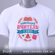 Футболка вчитель у світі білого кольору, складена на поверхні, фон світло-сірий, принт з текстом 'НАЙКРАЩИЙ ВЧИТЕЛЬ У СВІТІ' у рожевих та блакитних тонах, з елементами, такими як книга, дзвоник та ручка, з етикеткою на комірці.