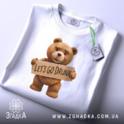 Футболка ведмедик let’s drunk, біла, складена на столі, фон світло-сірий, принт з ведмедиком, який тримає табличку з написом 'LET'S GO DRUNK', на комірці є етикетка.