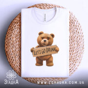 Футболка ведмедик let’s drunk, біла, складена на плетеній основі, фон світло-сірий, принт з ведмедиком, який тримає табличку з написом 'LET'S GO DRUNK'.