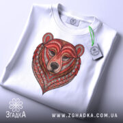 Футболка ведмідь білого кольору, складена на столі, фон світлий, принт ведмедя з червоними та коричневими деталями, на комірці прикріплена етикетка зеленого кольору.