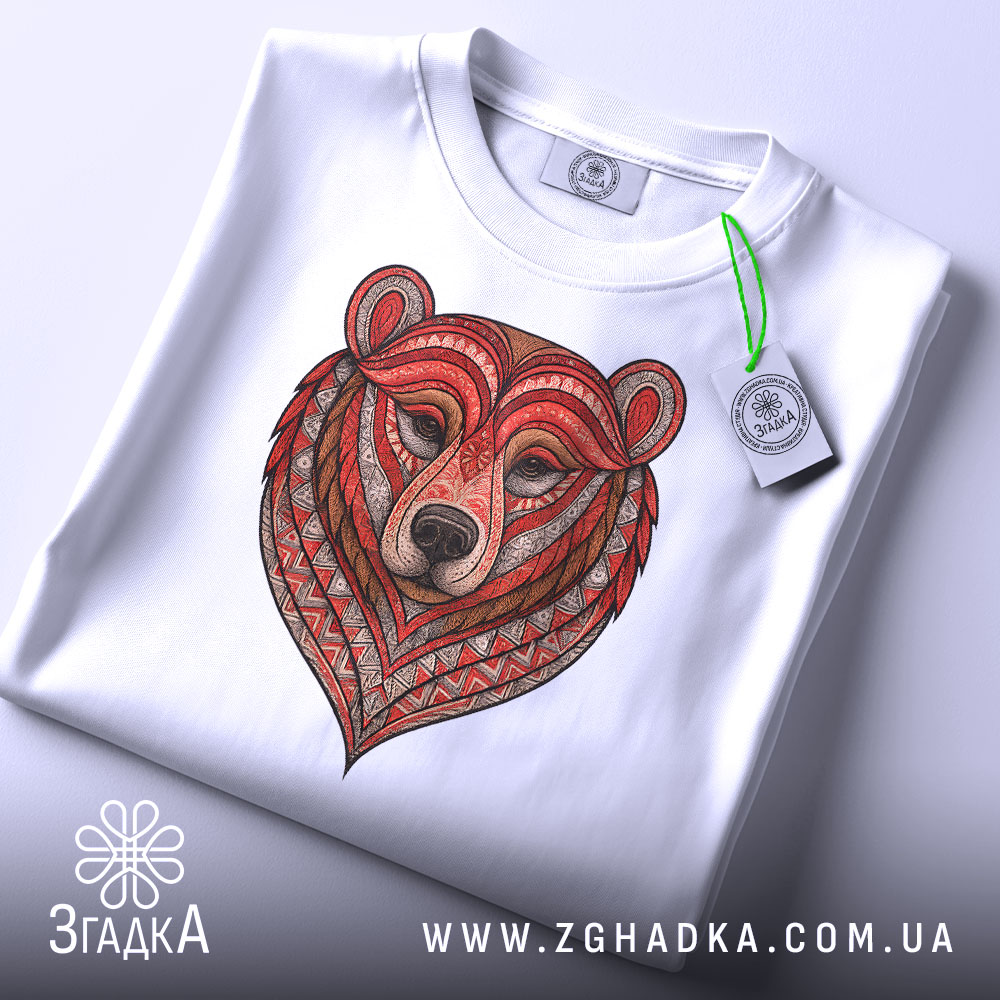 Футболка ведмідь білого кольору, складена на столі, фон світлий, принт ведмедя з червоними та коричневими деталями, на комірці прикріплена етикетка зеленого кольору.