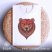 Футболка ведмідь білого кольору, складена на плетеній кошику, фон світлий, принт ведмедя з червоними та коричневими деталями, оформлений у стилі геометричних візерунків.