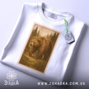 Футболка ведмідь білого кольору, складена на поверхні, з видимим етикеткою. Принт з ведмедем в лісі в коричневих відтінках, обрамлений рамкою, що нагадує поштову марку, на фоні світло-сірого градієнту.