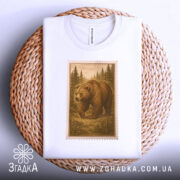 Футболка ведмідь білого кольору, складена на плетеній кошику, з видимим принтом ведмедя в лісі в коричневих відтінках, обрамленим рамкою, що нагадує поштову марку, на фоні світло-сірого градієнту.