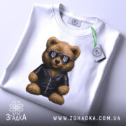 Футболка ведмідь патріот, біла, складена на поверхні, фон світло-сірий, принт ведмедя в чорній куртці та сонцезахисних окулярах з тризубом, з етикеткою.