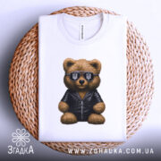 Футболка ведмідь патріот, біла, складена на плетеній кошику, фон світло-сірий, принт ведмедя в чорній куртці та сонцезахисних окулярах з тризубом.