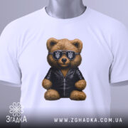 Футболка ведмідь патріот, біла, складена на поверхні, фон світло-сірий, принт ведмедя в чорній куртці та сонцезахисних окулярах з тризубом, з етикеткою.