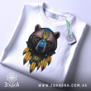 Футболка ведмідь символ, біла, складена на столі, фон світло-сірий, принт ведмедя з українським гербом, на комірці є етикетка.