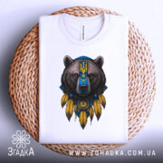 Футболка ведмідь символ, біла, складена на плетеній кошику, фон світло-сірий, принт ведмедя з українським гербом та жовтими пір'ями.