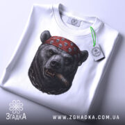 Футболка ведмідь з сигарою, біла, складена, зображення ведмедя в червоній бандані з черепами, ракурс зверху, фон світлий, на етикетці зелена стрічка.