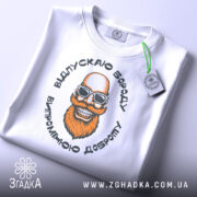 Футболка відпускаю бороду, білого кольору, лежить на плоскій поверхні, фон світло-сірий, принт з усміхненим бородатим чоловіком у сонцезахисних окулярах, на футболці є етикетка з зеленим шнурком.