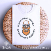 Футболка відпускаю бороду, білого кольору, лежить на плетеній кошику, фон світло-сірий, принт з усміхненим бородатим чоловіком у сонцезахисних окулярах, видно етикетку на горловині.