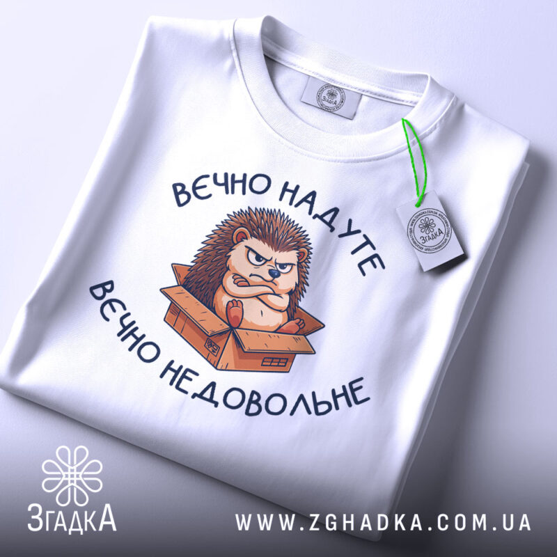 Футболка вєчно недовольне, білого кольору, зображена зверху, на ній видно принт з їжаком в коробці, фон світло-сірий, на комірці є етикетка.