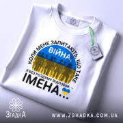 Футболка Війна це імена. Біла футболка, складена на столі, з видимим етикеткою. Принт на футболці містить текст 'ВІЙНА' і силуети людей на фоні жовто-блакитного прапора. Фон світлий, з легким відблиском.