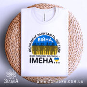 Футболка Війна це імена. Біла футболка, складена на плетеному кошику. Принт містить текст 'ВІЙНА' і силуети людей на фоні жовто-блакитного прапора. Фон світлий, з натуральними текстурами.