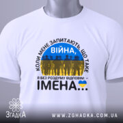 Футболка Війна це імена. Біла футболка, складена на столі, з видимим етикеткою. Принт на футболці містить текст 'ВІЙНА' і силуети людей на фоні жовто-блакитного прапора. Фон світлий, з легким відблиском.