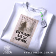 Футболка він полює відчуває, біла, зображена зверху, на фоні світлого градієнту, принт з текстом "ВІН ПОЛЮЄ. АЛЕ ЩЕ ВІДЧУВАЄ." на старовинному фоні, з зеленим ярликом.