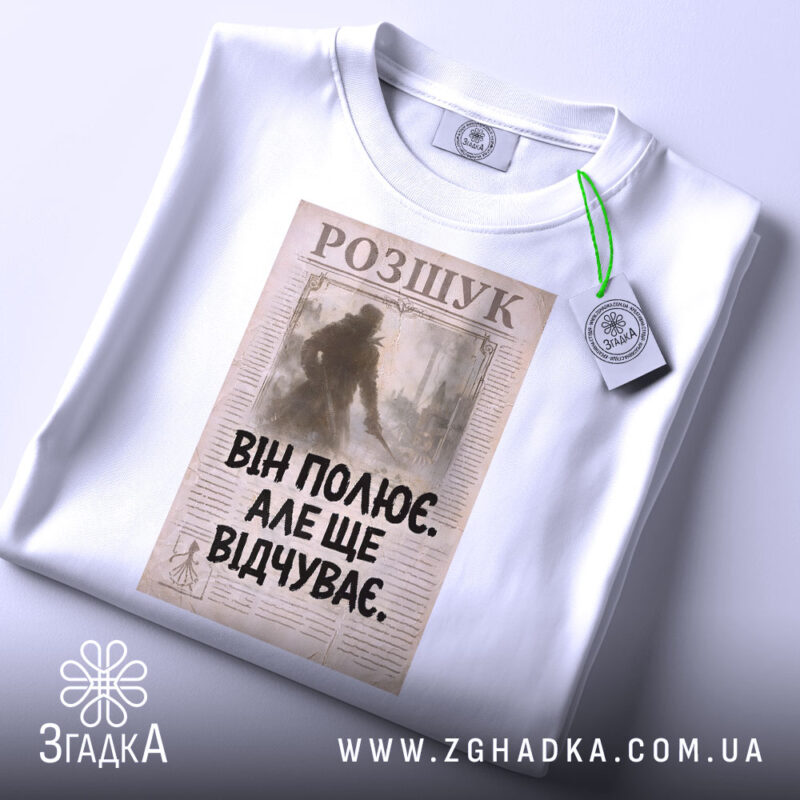 Футболка він полює відчуває, біла, зображена зверху, на фоні світлого градієнту, принт з текстом "ВІН ПОЛЮЄ. АЛЕ ЩЕ ВІДЧУВАЄ." на старовинному фоні, з зеленим ярликом.