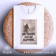 Футболка він полює відчуває, біла, зображена зверху, на фоні плетеного кошика, принт з текстом "ВІН ПОЛЮЄ. АЛЕ ЩЕ ВІДЧУВАЄ." на старовинному фоні.