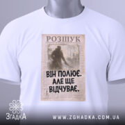 Футболка він полює відчуває, біла, зображена зверху, на фоні світлого градієнту, принт з текстом "ВІН ПОЛЮЄ. АЛЕ ЩЕ ВІДЧУВАЄ." на старовинному фоні, з зеленим ярликом.