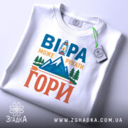 Футболка віра може рухати гори. Біла футболка, складена на плоскій поверхні, з видимим етикеткою. На фоні світло-сірий градієнт. Принт на футболці повторює текст 'ВІРА МОЖЕ РУХАТИ ГОРИ' з малюнком ліхтаря та гір, виконаним у яскравих кольорах.