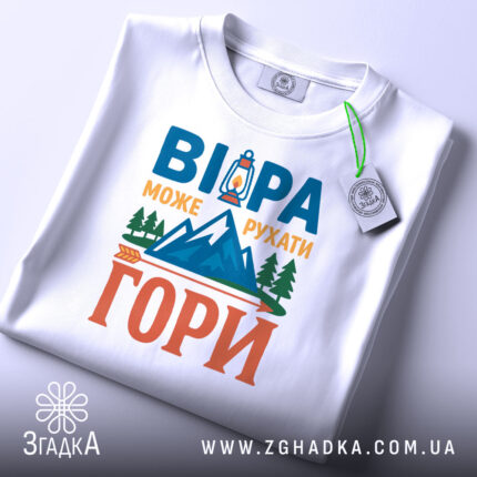 Футболка віра може рухати гори. Біла футболка, складена на плоскій поверхні, з видимим етикеткою. На фоні світло-сірий градієнт. Принт на футболці повторює текст 'ВІРА МОЖЕ РУХАТИ ГОРИ' з малюнком ліхтаря та гір, виконаним у яскравих кольорах.