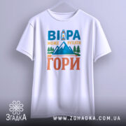Футболка віра може рухати гори. Біла футболка з коротким рукавом, підвішена на вішалці. На фоні світло-сірий градієнт. Принт містить текст 'ВІРА МОЖЕ РУХАТИ ГОРИ' з малюнком гір і ліхтаря у яскравих кольорах.