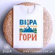 Футболка віра може рухати гори. Біла футболка, складена на плоскій поверхні, розташована на плетеній основі. На фоні світло-сірий градієнт. Принт містить текст 'ВІРА МОЖЕ РУХАТИ ГОРИ' з малюнком ліхтаря та гір у яскравих кольорах.