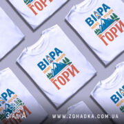 Футболка віра може рухати гори. Біла футболка з коротким рукавом, зображена на манекені з металевим блиском. На фоні світло-сірий градієнт. Принт містить текст 'ВІРА МОЖЕ РУХАТИ ГОРИ' у яскравих кольорах: синьому, оранжевому та зеленому, з малюнком гір і ліхтаря.