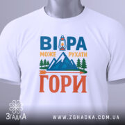 Футболка віра може рухати гори. Біла футболка, складена на плоскій поверхні, з видимим етикеткою. На фоні світло-сірий градієнт. Принт на футболці повторює текст 'ВІРА МОЖЕ РУХАТИ ГОРИ' з малюнком ліхтаря та гір, виконаним у яскравих кольорах.