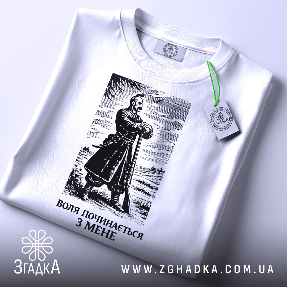 Футболка воля починається з мене. Біла футболка з коротким рукавом, зображена з верхнього ракурсу на фоні світлого кольору. На футболці чорний принт з зображенням чоловіка в одязі 19 століття, а також видна етикетка з зеленим шнурком. Текст під зображенням: 'ВОЛЯ ПОЧИНАЄТЬСЯ З МЕНЕ'.