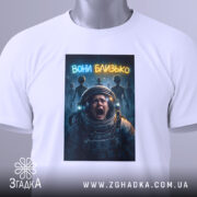 Футболка вони близько, біла, зображена на столі, фон світло-сірий, принт з космонавтом, який кричить, на фоні силуети прибульців і написом 'вони близько', з етикеткою на шнурку.