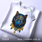 Футболка вовк білого кольору, складена на поверхні, з принтом вовка в синіх та жовтих тонах, вид з верхнього ракурсу, фон світло-сірий, на футболці є етикетка з зеленим шнурком.