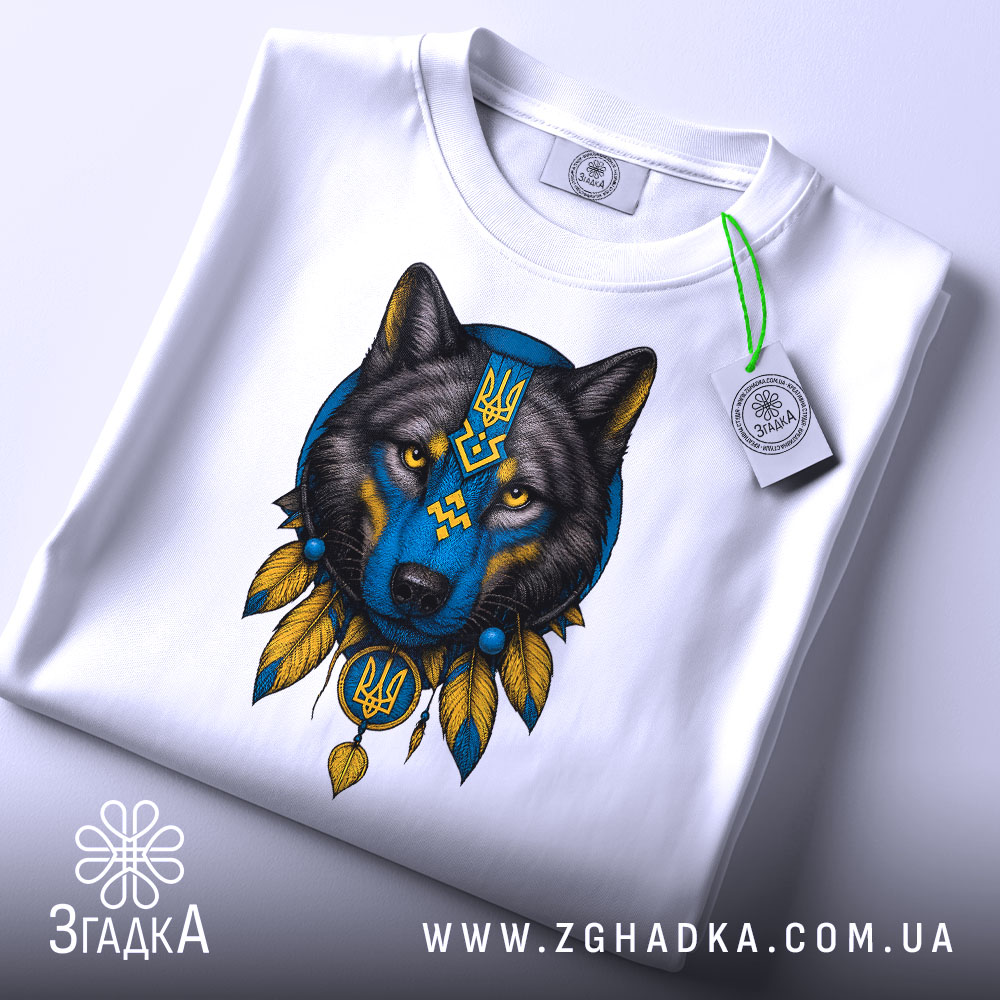 Футболка вовк білого кольору, складена на поверхні, з принтом вовка в синіх та жовтих тонах, вид з верхнього ракурсу, фон світло-сірий, на футболці є етикетка з зеленим шнурком.