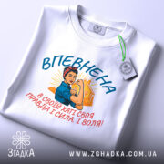 Футболка впевнена сильна жінка, білого кольору, зображена зверху, фон світло-сірий, принт з жінкою в синій блузці та червоній пов'язці, текст на українській мові, прикріплена етикетка з логотипом.