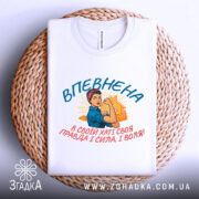 Футболка впевнена сильна жінка, білого кольору, зображена на плоскій поверхні, фон світло-сірий, принт з жінкою в синій блузці та червоній пов'язці, текст на українській мові, розташована на плетеній основі.
