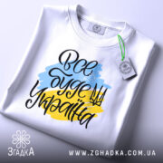 Футболка все буде Україна, біла, зображена зверху, на столі, фон світло-сірий, принт з написом 'Все буде Україна' у жовто-блакитних кольорах, з етикеткою на зеленій стрічці.