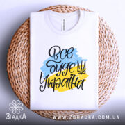 Футболка все буде Україна, біла, зображена зверху на плетеній основі, фон світло-сірий, принт з написом 'Все буде Україна' у жовто-блакитних кольорах.