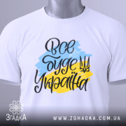 Футболка все буде Україна, біла, зображена зверху, на столі, фон світло-сірий, принт з написом 'Все буде Україна' у жовто-блакитних кольорах, з етикеткою на зеленій стрічці.